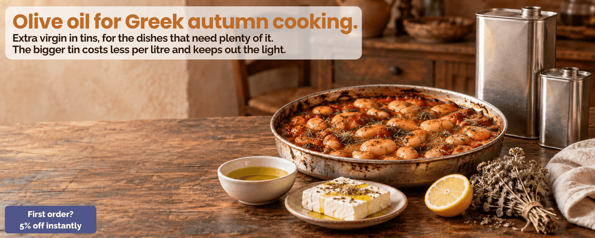 Two tins of Greek extra virgin olive oil beside a baking pan of gigantes plaki, feta with oregano and a bowl of olive oil on a wooden table