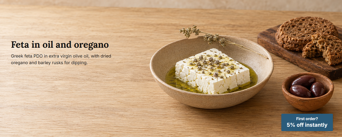 Feta in a shallow bowl of olive oil with crumbled dried oregano, barley rusks and olives on an oak table