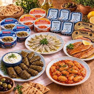 Griechische Meze-Vorratsbox: Dolmadakia, Gigantes, Sardellen mit Oregano und Räucherhering auf Tellern, mit den Dosen von Zanae und Trata auf einem Holztisch