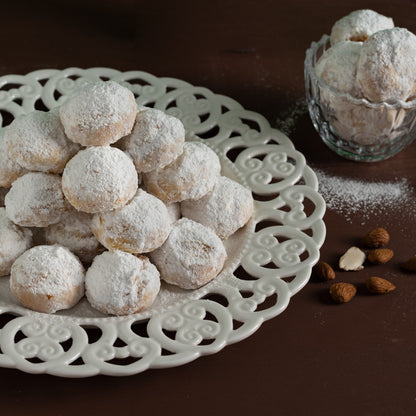 Kourabiedes sur une assiette blanche ajourée avec des amandes et du sucre glace, sur une table en bois sombre