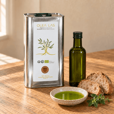 Silberne 3-Liter-Dose Olea Las Olivenöl mit einer Glasflasche, geschnittenem Brot, einer Schale Olivenöl und frischem Oregano auf einem Eichentisch