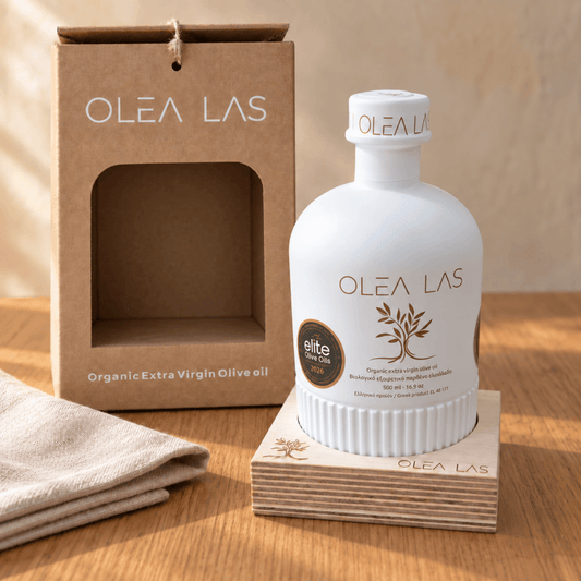 Botella blanca de 500 ml de aceite de oliva Olea Las sobre su base de madera grabada, junto a la caja regalo de cartón kraft abierta, en una mesa de roble