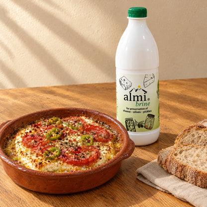 Ofen-Feta mit Tomate, Chili und Olivenöl in einer Tonform, dahinter die Flasche Almi-Salzlake