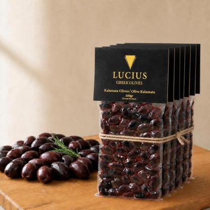 Six Lucius Kalamata olive pouches of 250g tied with twine beside a pile of olives on an oak board